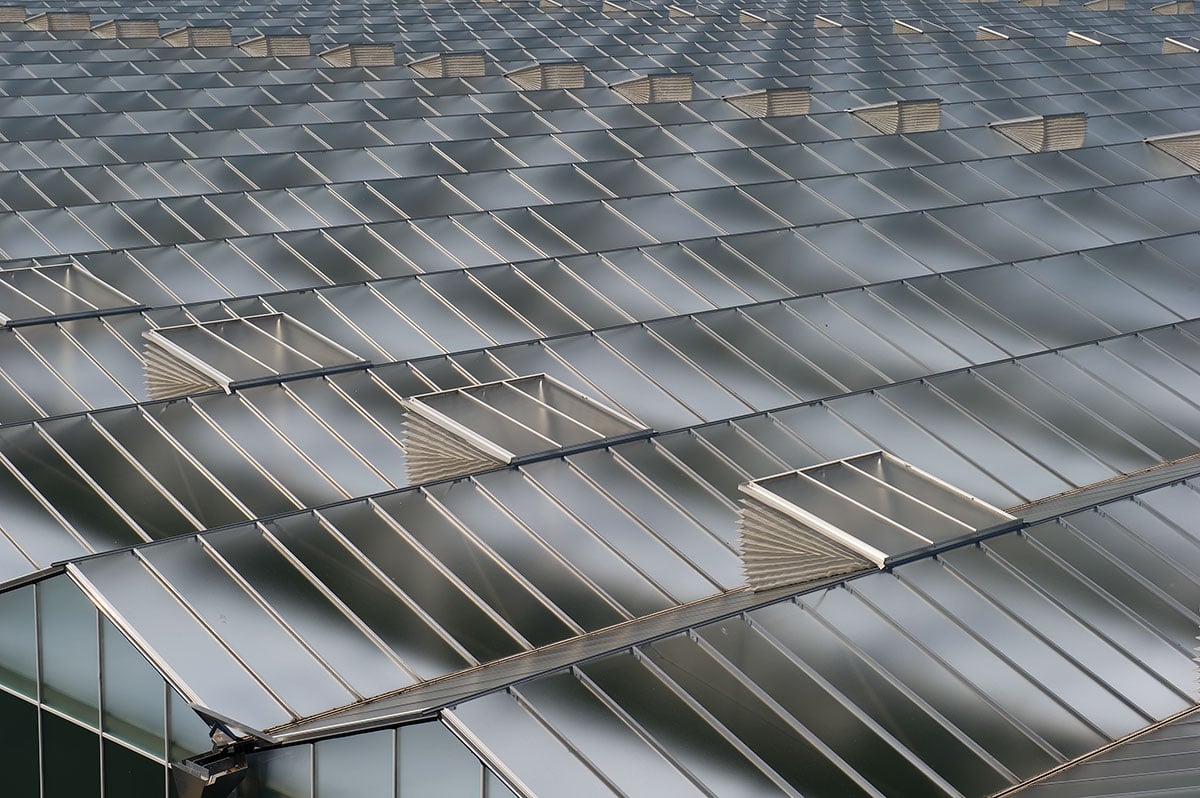 The image shows a roof of a modern greenhouse. The roof has a reflective surface, creating a shimmering effect. Between some of the glass windows, there are openings or vents. The angle of the image is from below, looking up towards the top of the building.