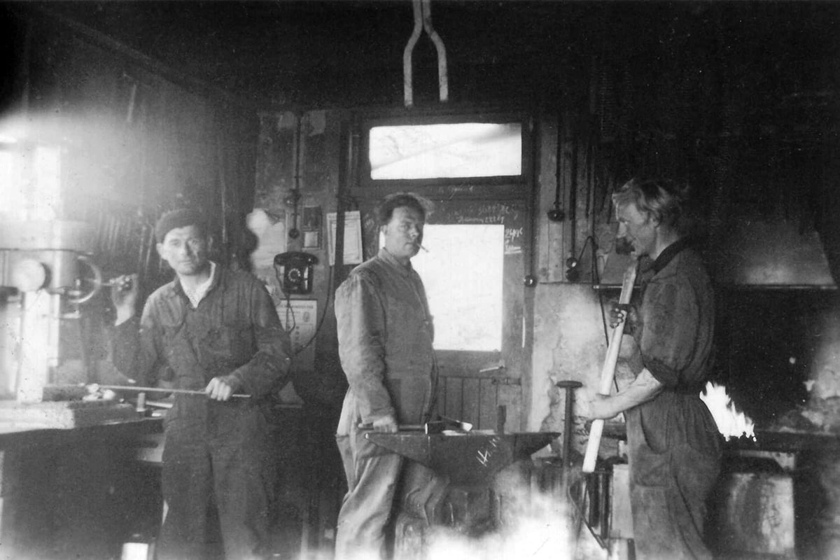 A black and white photograph of four individuals in an early 20th-century workshop setting. They are dressed in period-appropriate work attire, including overalls and hats, and appear to be engaged in a task that involves machinery or equipment.