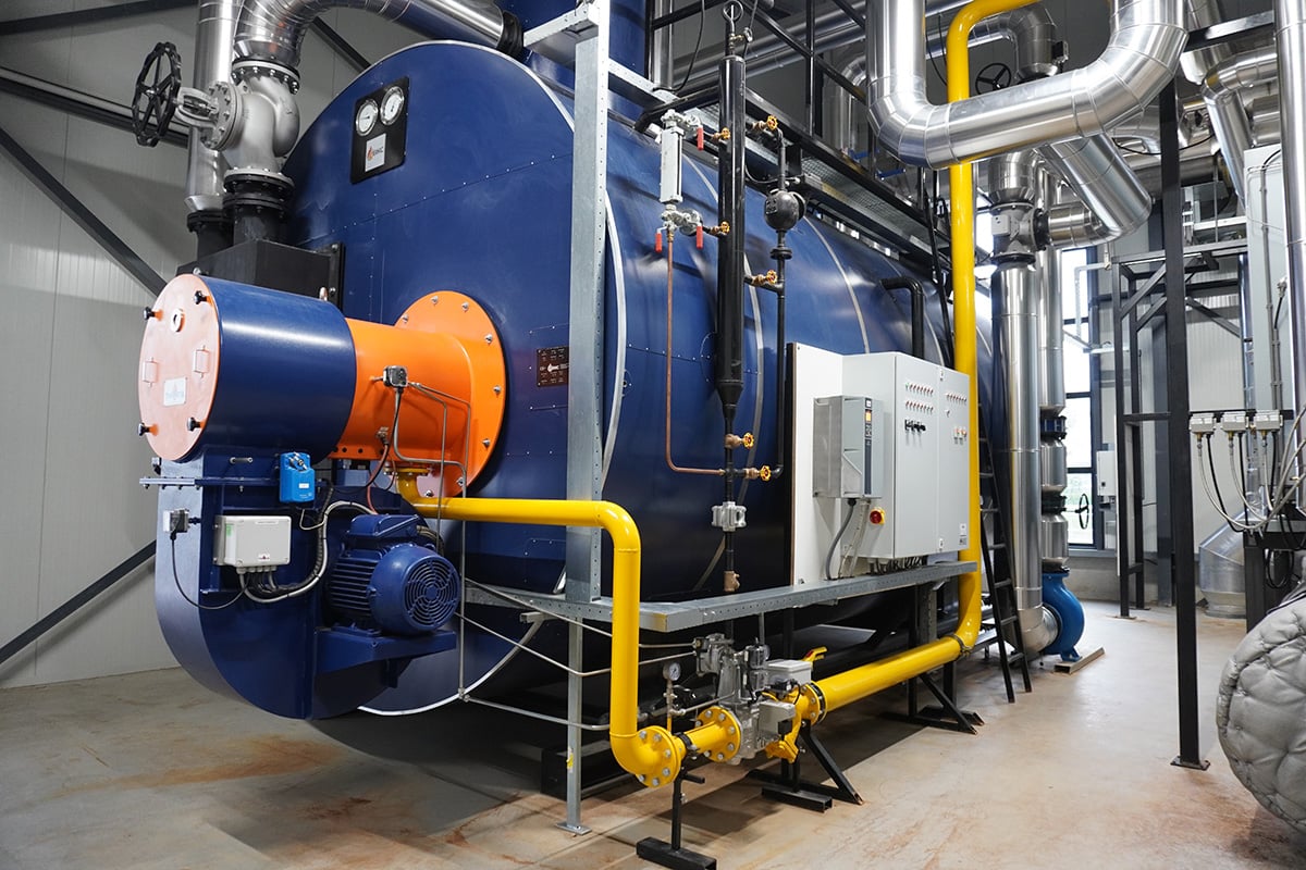 An industrial setting with a large blue steam boiler as the central focus. The boiler is equipped with an orange safety valve on top, and there are multiple pipes and valves around it, suggesting it is part of a larger heating or power generation system.